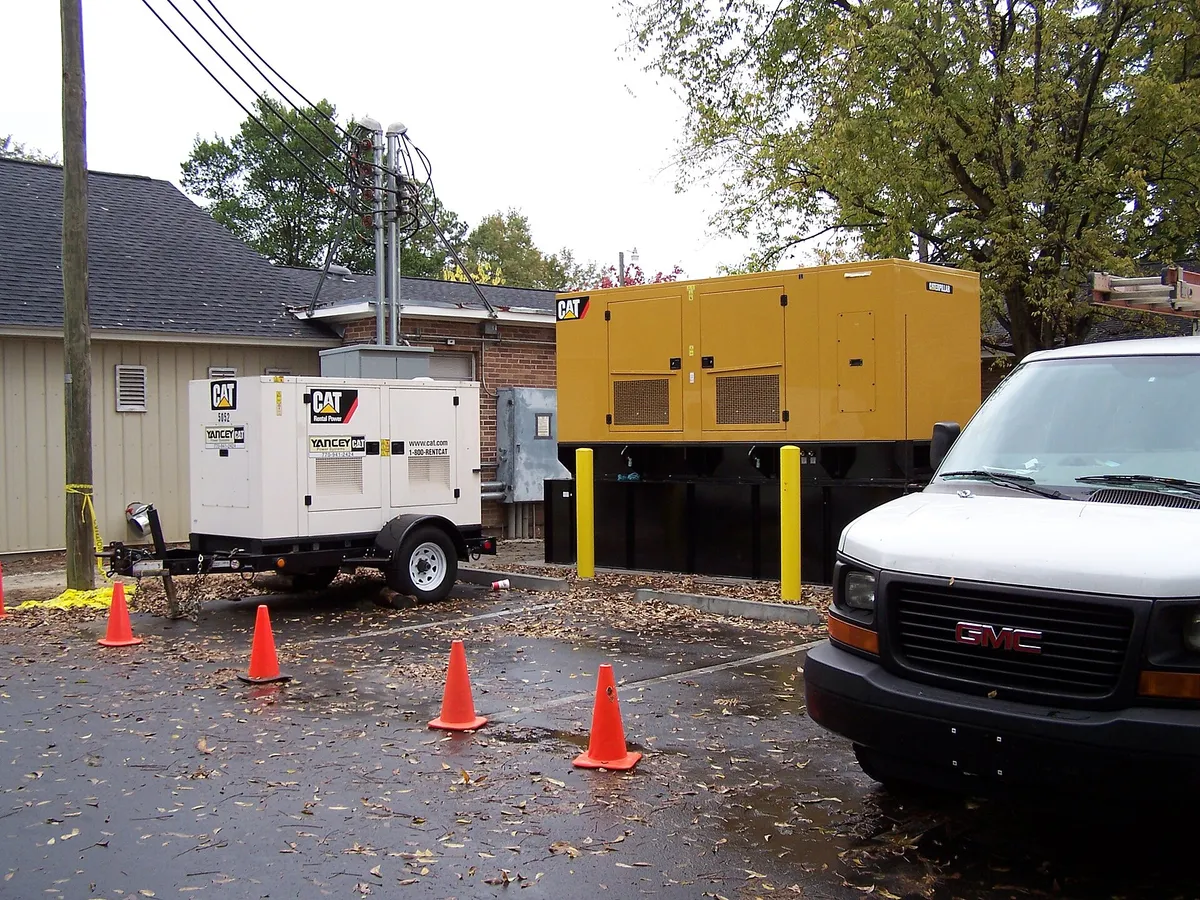 Genset portabel putih di atas trailer beroda di sebelah genset berkanopi kuning yang jauh lebih besar, dengan kerucut lalu lintas di depannya