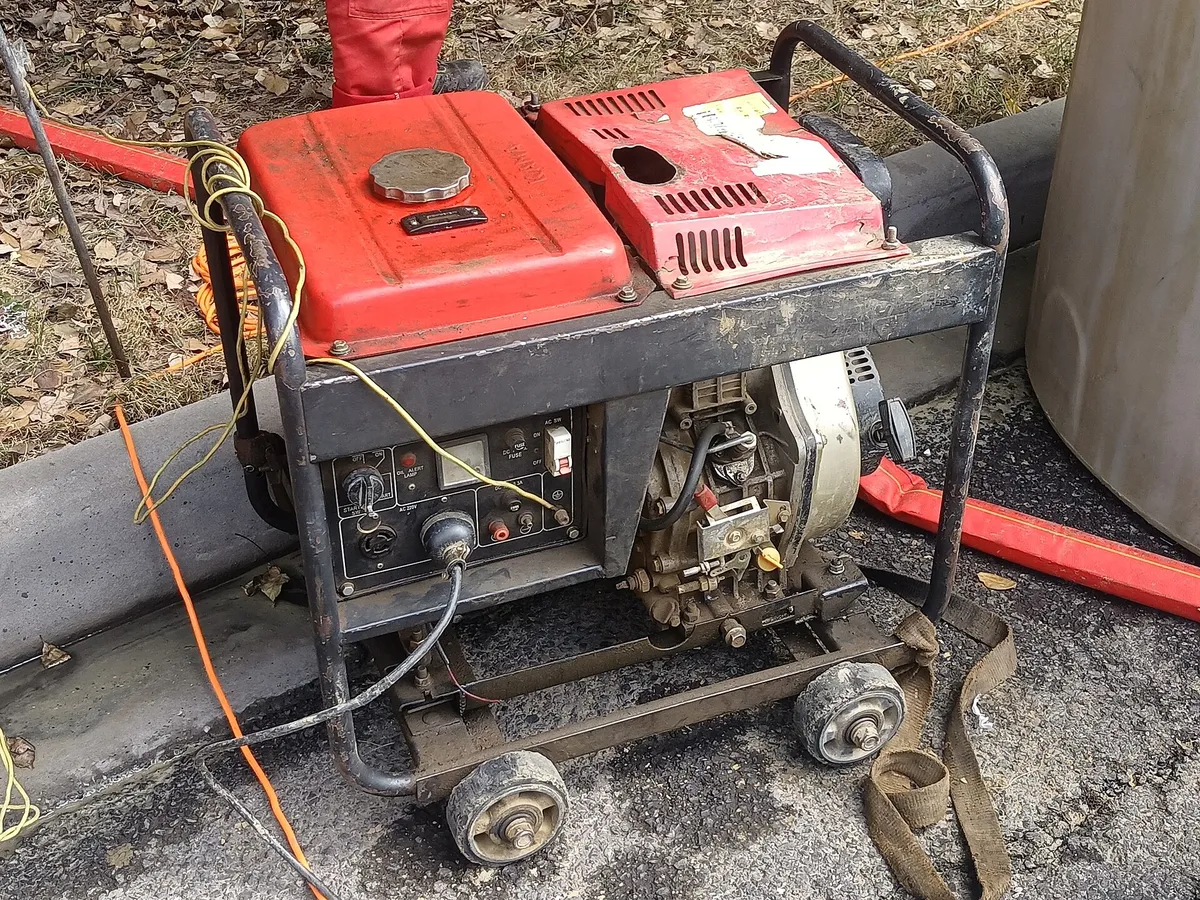 Genset diesel portabel kecil beroda dengan panel kontrol di sisi mesin, berdiri di atas aspal dengan kabel menjulur