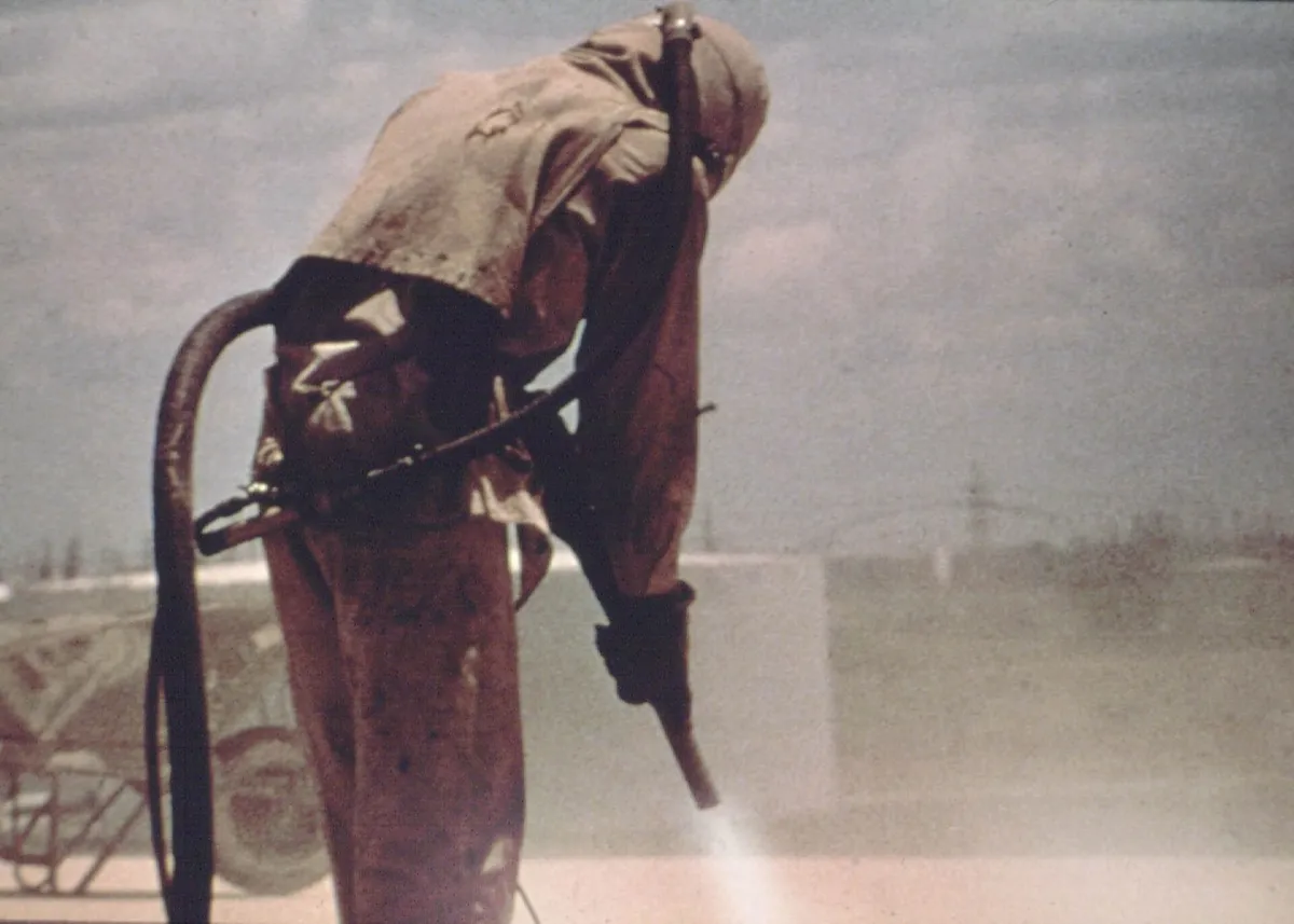 Operator sandblasting memakai helm dan pakaian pelindung penuh sambil mengarahkan nozzle ke permukaan