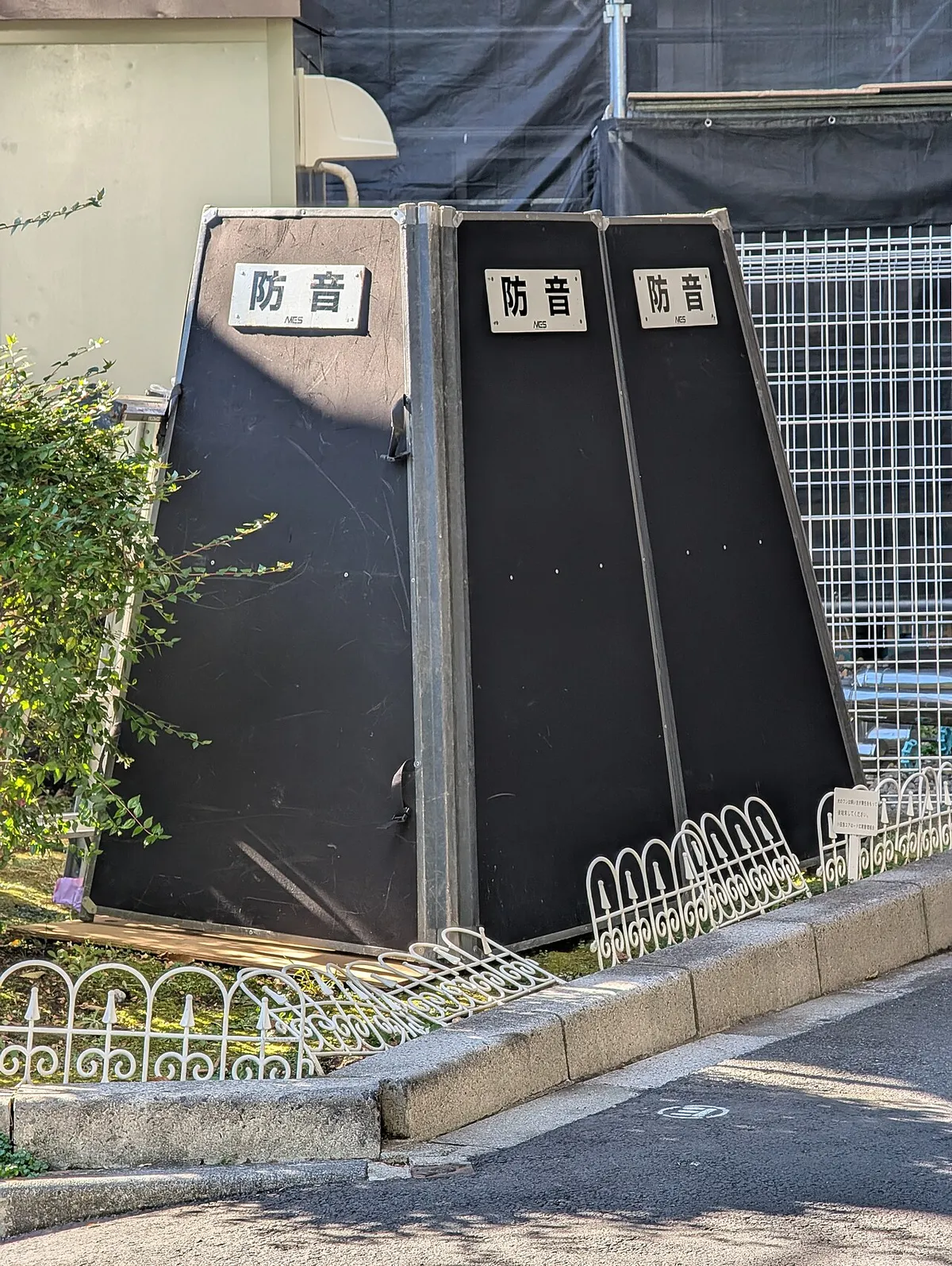 Tiga panel peredam suara hitam bertanda huruf Jepang bōon (peredam suara) berdiri di tepi jalan di depan pagar